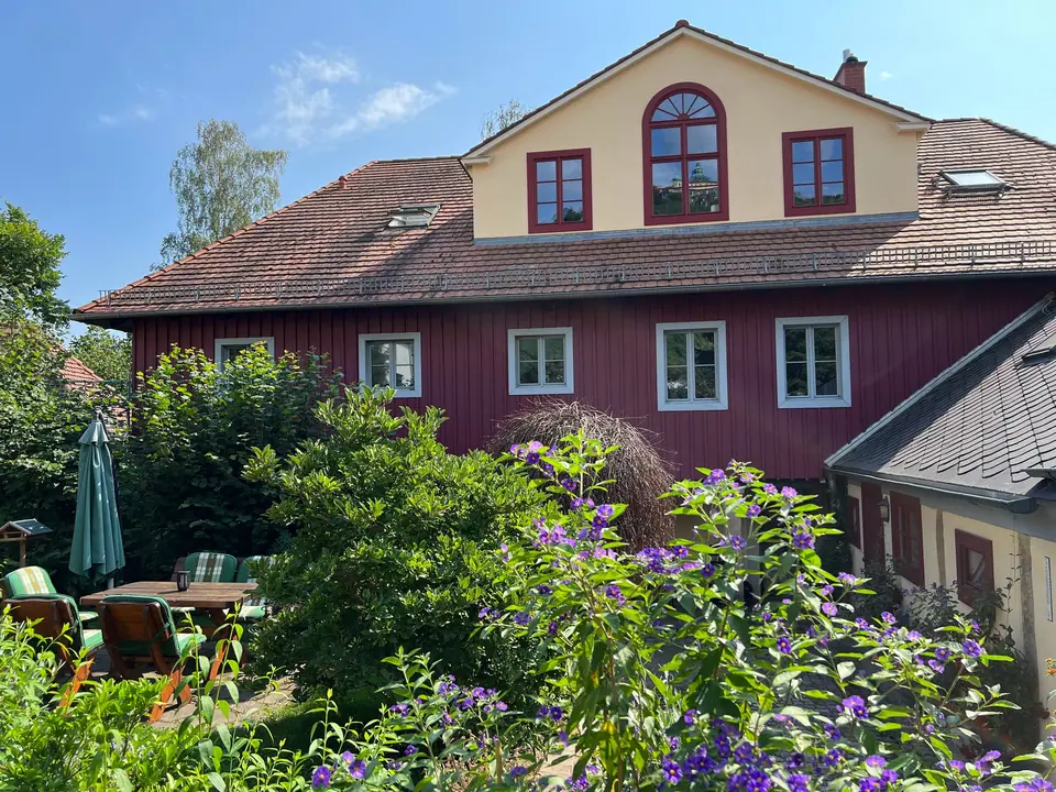 Die Gartenseite des Wohnhauses mit Sitzplatz, blühenden Sträuchern und dem ochsenblutroten Obergeschoss